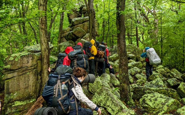a group of students wearing backpacks make their way uphill in a green forest 